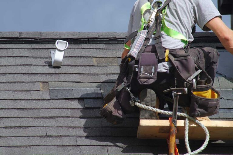 Homeowner placing a bucket beneath a ceiling leak while storm-damaged roofing is visible through a window.
